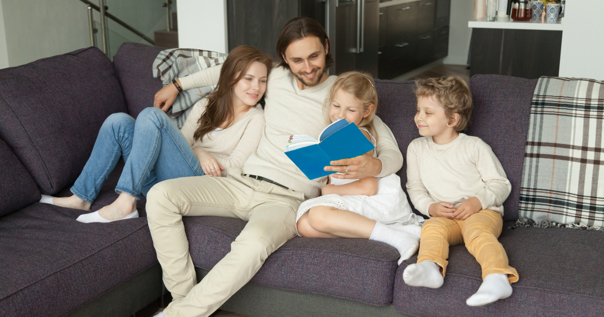 A happy family sitting together in a bright living room, parents with two children, laughing or reading together