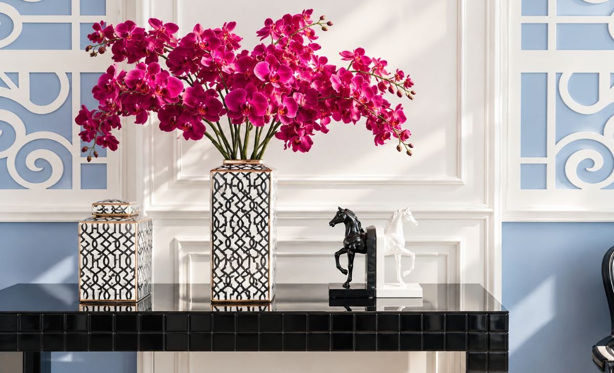 A sophisticated interior decor arrangement featuring vibrant pink orchids in a patterned vase, matching decorative accessories, black and white horse sculptures, and elegant blue and white wall panels above a glossy black console table.